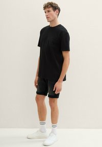 Czarna koszulka z krótkim rękawem i kieszonką, zestawiona z ciemnymi szortami dżinsowymi. Noszona z białymi trampkami i paskowanymi skarpetkami do kostek. Gładka faktura.