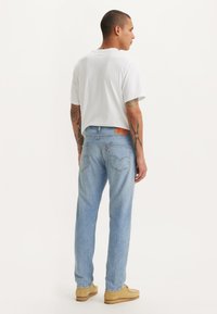 Lichtblauwe jeans met een rechte pasvorm, met een leren label op de achterkant, gedragen met een eenvoudig wit t-shirt en beige schoeisel.