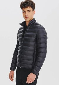 Czarna kurtka puchowa z pikowanym wzorem, wysokim kołnierzem i zapięciem na zamek. Zawiera akcent z logo na lewym rękawie.