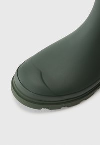 Botte en caoutchouc verte avec un embout rond, texture lisse et semelle robuste, présentant des détails subtils sur le bord et des éléments de traction.
