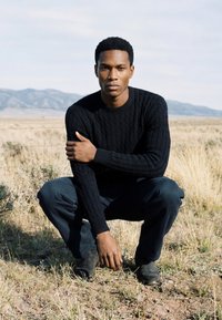 Homme portant un pull noir et un pantalon sombre accroupi dans un champ herbeux avec des montagnes en arrière-plan sous un ciel clair.