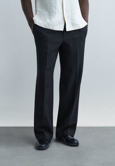 Personne debout les mains dans les poches portant un pantalon noir sur mesure, des chaussures habillées noires et une chemise blanche à manches courtes sur un fond uni.