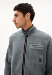 Giacca in pile grigia con collo alto, chiusura frontale con zip e tasca sul petto con un piccolo logo. Materiale texturizzato e caldo.