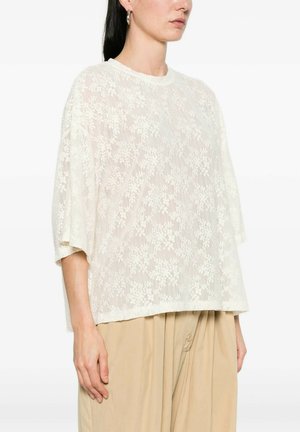 Donna che indossa una maglietta a maniche corte ampia bianca in pizzo floreale abbinata a pantaloni beige a vita alta con dettagli a bottoni.