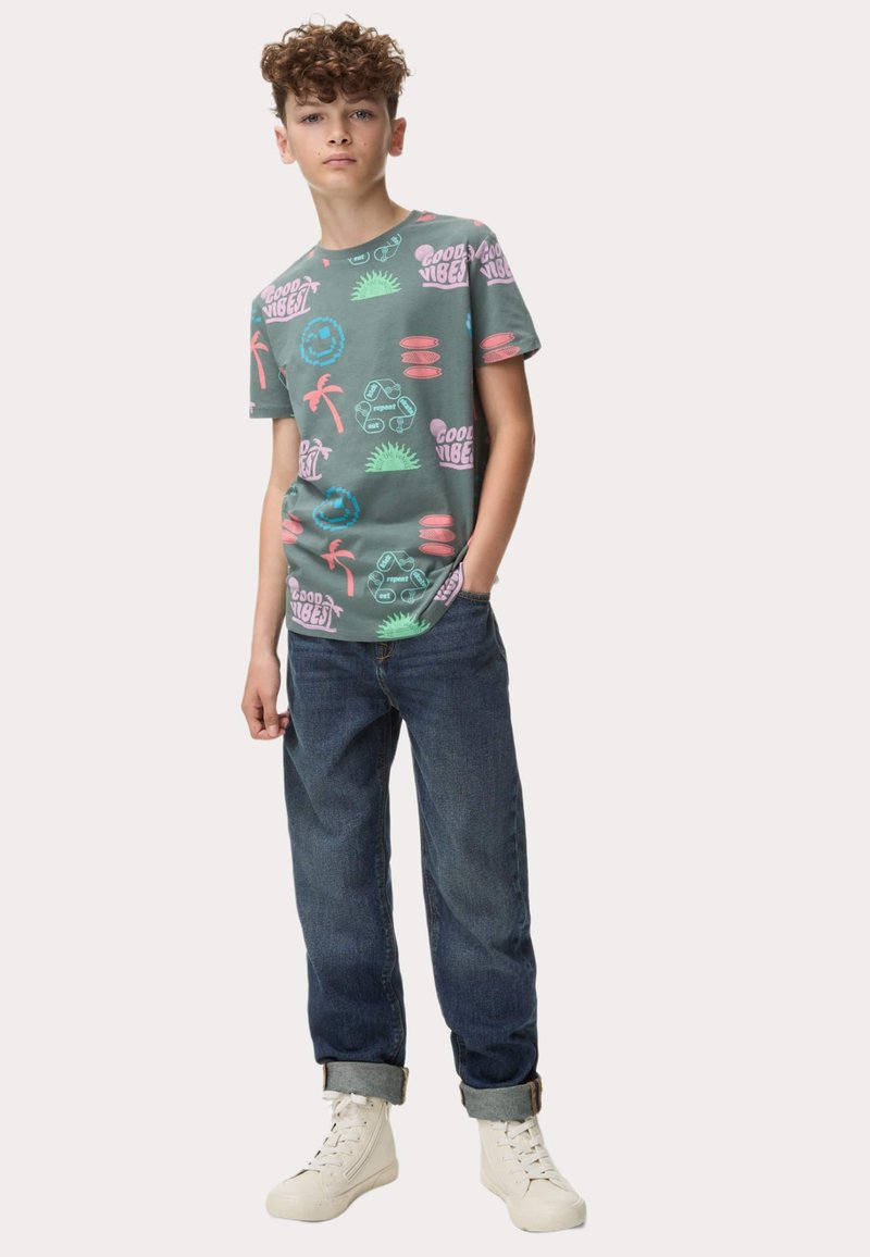 Camiseta de algodón verde con un estampado colorido de temática playera, con palmeras y texto. Combinada con jeans de denim oscuro arremangados y zapatillas blancas.