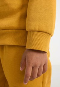 Senfgelbes Sweatshirt mit gerippten Bündchen und Saum, das eine glatte, weiche Textur aufweist. Eine Hand ruht auf dem Stoff.