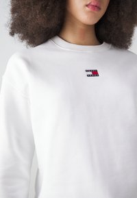 Biely sveter s okrúhlym výstrihom, ktorý má na prednej strane malú, obdĺžnikovú, viacfarebnú nášivku s nápisom "TOMMY JEANS". Mäkká textúra.