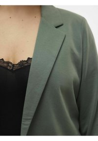 Blazer vert avec un col châle, fabriqué en tissu doux. Porté sur un top noir bordé de dentelle, montrant une texture lisse et un design superposé.