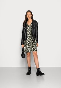 Jaqueta de motoqueiro de couro preto sobre um vestido estampado em verde e preto, combinada com botas de tornozelo pretas e uma pequena bolsa trançada.