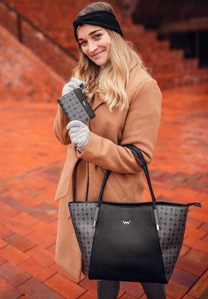 Black and gray tote bag with patterned side, featuring a central logo and sturdy handles. Model holds a matching wallet. Brown coat.