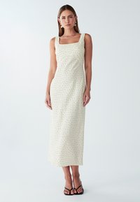 Maxikleid aus cremefarbenem Stoff mit schwarzen Punkten, quadratischem Ausschnitt, breiten Trägern und einer figurbetonten Silhouette. Getragen mit schwarzen, schnürbaren Sandalen.