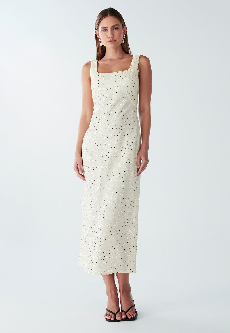 Maxikleid aus cremefarbenem Stoff mit schwarzen Punkten, quadratischem Ausschnitt, breiten Trägern und einer figurbetonten Silhouette. Getragen mit schwarzen, schnürbaren Sandalen.