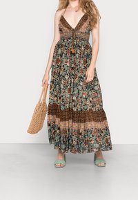 Maxi sukienka w kwiatowe wzory w ciemnych odcieniach, z głębokim dekoltem w serek, falbaniastymi ramiączkami i warstwową spódnicą. Noszona z zielonymi sandałami na słupku.