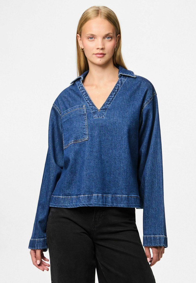 Tumši zila džinsa krekla blūze ar V veida kaklu, garām piedurknēm, kabatas uzlīmi un saīsinātu apakšu. Mīksta tekstūra ar redzamām šuvēm.