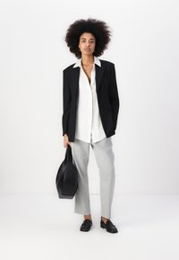 Veste noire ajustée, chemise blanche, pantalon gris clair, mocassins noirs et un sac en cuir noir. Textures lisses, design structuré.