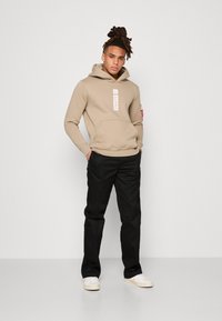 Beige hettegenser med logo-patch, frontlomme og justerbar snor. Kombinert med svarte bukser og hvite sneakers. Glatt tekstur.