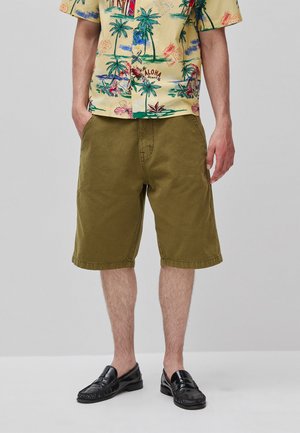 Mann trägt olivgrüne knielange Shorts, schwarze Loafers und ein gelbes Hawaii-Hemd mit Palmen- und Blumenmuster.