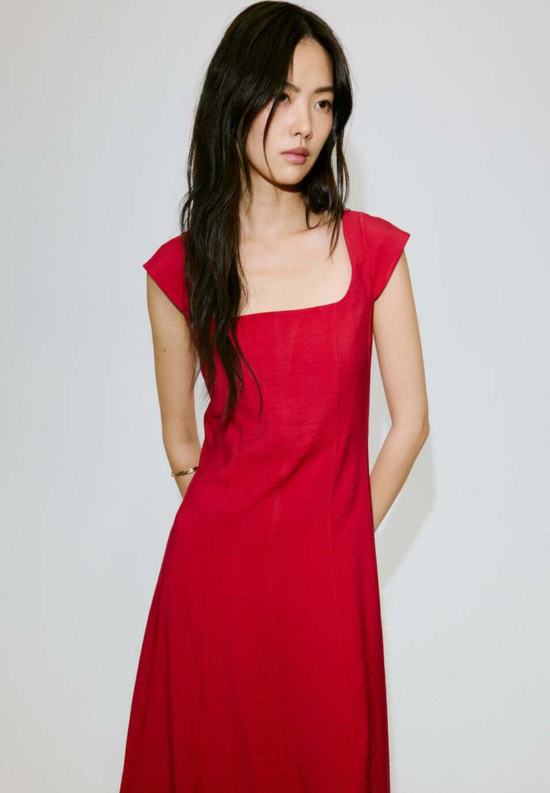 Femme aux longs cheveux foncés portant une robe rouge à manches courtes et un décolleté carré, debout devant un fond clair uni.