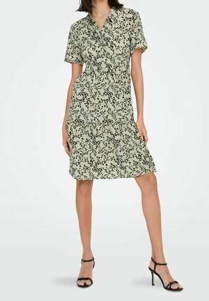 Femme portant une robe vert clair longueur genou avec motif de points noirs irréguliers, manches courtes, col et sandales noires à talons hauts.