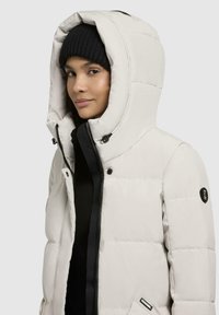 Lett grå puffjakke med avtagbar hette, svart glidelås og logo-patch. Har en quiltet design og en myk, glatt tekstur.