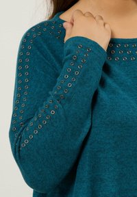 Maglione turchese con una finitura testurizzata, caratterizzato da occhielli in metallo silver lungo le maniche, creando un design di accento unico.