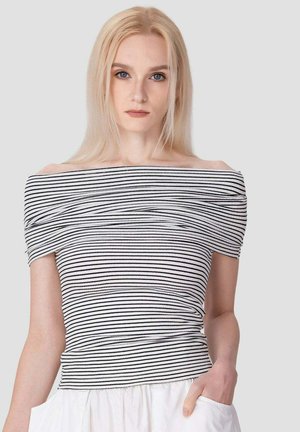 Jeune femme aux longs cheveux blonds porte un haut rayé noir et blanc épaules dénudées et une jupe blanche, main dans la poche, fond neutre.
