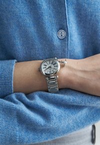 Srebrny zegarek na nadgarstek ze stali nierdzewnej z białą tarczą, czarnymi rzymskimi cyframi i czerwonym akcentem. Noszony na jasnoniebieskim dzianinowym swetrze.