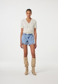 Romantische kortemouwen cardigan in roomkleur met bloemenprint en gouden knopen, gecombineerd met blauwe denim shorts en beige kniehoge laarzen met studs.