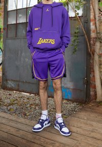 Violetti huppari, jossa on "Lakers"-logo ja vetoketjullinen tasku, yhdistettynä violettiin shortsiin, jossa on keltaisia yksityiskohtia. Valkoiset ja violetit lenkkarit täydentävät asukokonaisuutta.
