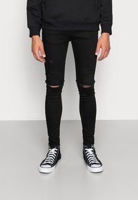 Jeans negros ajustados con rodillas rasgadas, combinados con zapatillas altas negras con cordones blancos y suelas de goma. Textura suave, ajuste casual.