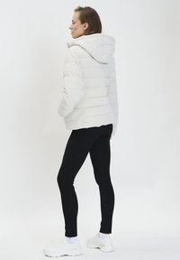 Giacca bianca gonfiabile con cappuccio, caratterizzata da sezioni trapuntate orizzontali. Abbinata a leggings neri e scarpe sportive bianche. Sfondo neutro.