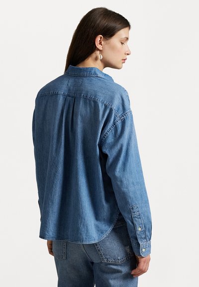 Cămașă din denim albastru deschis, cu închidere cu nasturi în față, mâneci lungi și un pliț în spate. Are un guler clasic și manșete cu nasturi.