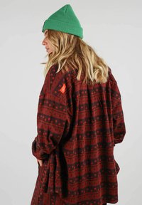 Chemise en flanelle oversized à manches longues, rouge et noir, avec des motifs géométriques. Elle comporte une poche et une étiquette en haut à gauche. Portée avec un bonnet vert.
