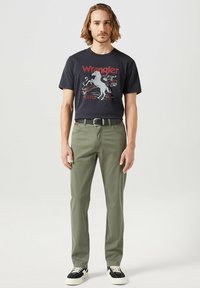 Mörk t-shirt med grafiskt tryck, olivgröna byxor, svart bälte och svarta sneakers med vita accenter, stående mot en neutral bakgrund.