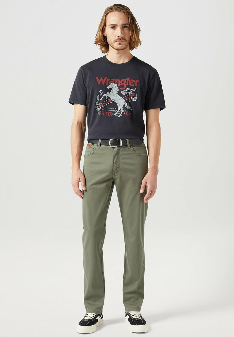 Mörk t-shirt med grafiskt tryck, olivgröna byxor, svart bälte och svarta sneakers med vita accenter, stående mot en neutral bakgrund.