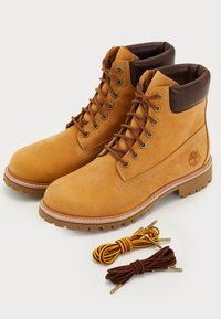 Paire de bottes en cuir tanné à lacets, avec des cols rembourrés brun foncé, logo embossé sur le côté, et deux jeux supplémentaires de lacets en dessous.