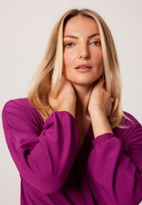 Fioletowy prążkowany top z obszernymi rękawami, z głębokim dekoltem. Modelka ma długie, blond włosy i pozuje, trzymając ręce na szyi.