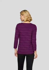 Lila Langarmshirt mit strukturierten, wellenförmigen Stoffschichten. Das Shirt hat einen runden Ausschnitt und wird mit schwarzen Hosen kombiniert.