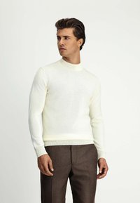Cremfarbener Strickpullover mit hohem Kragen, schlanker Passform und langen Ärmeln, kombiniert mit braunen, maßgeschneiderten Hosen. Glatte Textur, keine sichtbaren Muster.