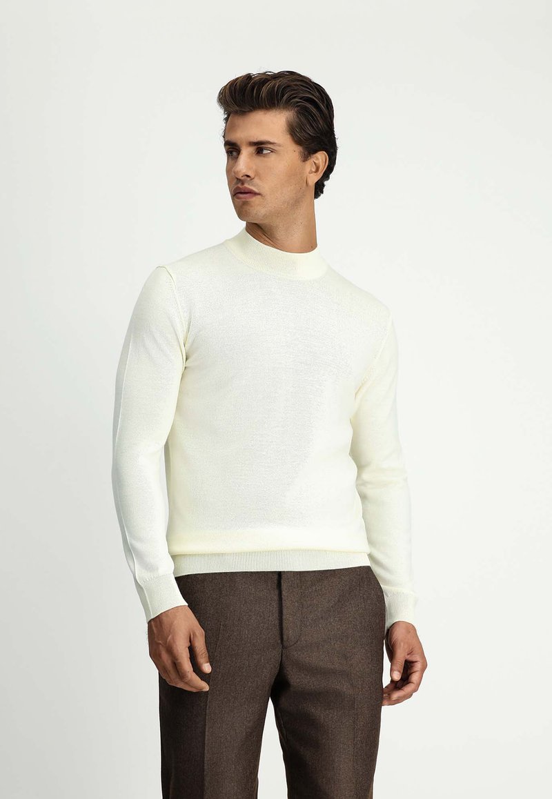 Cremfarbener Strickpullover mit hohem Kragen, schlanker Passform und langen Ärmeln, kombiniert mit braunen, maßgeschneiderten Hosen. Glatte Textur, keine sichtbaren Muster.