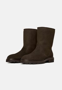 Botas de tobillo de ante marrón con punta redonda, suela sólida y parte superior texturizada. Presentan costuras mínimas y un diseño simple y robusto.