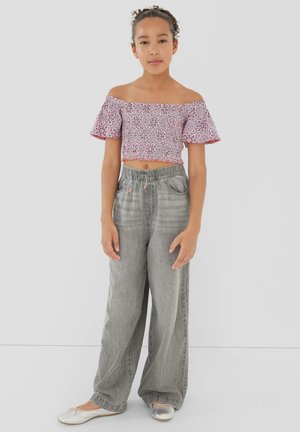 Jong meisje staand, draag een roze patroon off-shoulder crop top, losse grijze broek en zilveren ballerina's tegen een effen achtergrond.