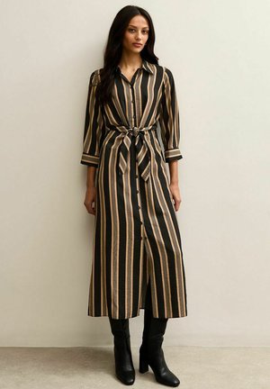 Frau trägt ein midi langes, schwarz-beiges, vertikal gestreiftes Hemdblusenkleid mit Gürtelknoten und schwarzen Stiefeln mit Absatz, steht vor einer schlichten Wand.