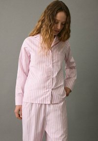 Mujer con pelo ondulado que lleva pijamas de botones a rayas blancas y rosa claro, de pie con una mano en el bolsillo, mirando hacia abajo.