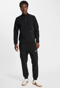 Zwarte fleecetrui met ritssluiting en hoge kraag, gecombineerd met bijpassende zwarte joggingbroek. Opvallend wit merklogo op de broek en subtiele accenten.