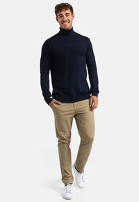 Mørkeblå turtleneck genser med teksturerte striper, kombinert med beige bukser og hvite sneakers. Modell som står med hendene i lommene.