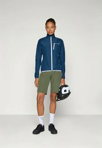 En person som har på seg en blå zip-up jakke, olivengrønne shorts, hvite sokker og svarte sko, holder en svart og hvit sykkelhjelm i den ene hånden.