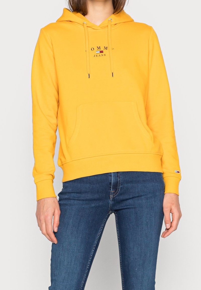 Gelbe Kapuzenjacke mit einer Bauchtasche, Kordelzug-Haube und "TOMMY JEANS"-Logo. Hergestellt aus weichem Stoff, mit gerippten Bündchen und Saum.