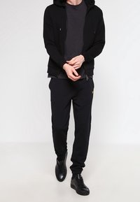 Czarna bluza z zamkiem i sznurkiem, szara koszulka pod spodem, czarne spodnie dresowe i czarne sneakersy. Prosty design bez wyraźnych wzorów.