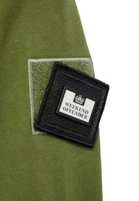 Olivgrüner Stoff mit einem quadratischen, schwarzen gewebten Patch, der den Text "WEEKEND OFFENDER" und ein Kronenlogo zeigt, vor einem weichen Hintergrund.
